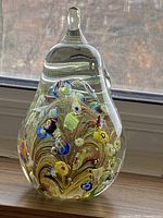 Front angled view of the pear-shaped paperweight showing colorful millefiori flowers and swirling golden patterns within clear glass.