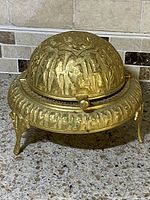 Closed view of vintage gold plated footed ornate hand etched caviar serving dish with rollback lid, showing detailed etched design on lid and base with feet.