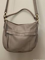 Full exterior front of the light beige/taupe leather shoulder bag showing the main zipper, front flap detail, and handle and shoulder strap attachments.