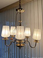 Full view of the 5-arm chandelier illuminated, showing the milk glass shades with gold leaf pattern and chrome frame with walnut accent.