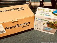Two boxes side by side. Larger box labeled AeroGarden Bounty, smaller box showing AeroGarden In-Home Garden System with image of flowering plants inside.
