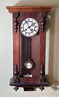 Front view of antique Vienna regulator wall clock, showing carved wooden case, white porcelain dial with Roman numerals, pendulum and finials.