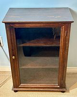 Front view of the wooden display cabinet showing glass door with brass knob, three interior shelves, and wood finish with minor wear.