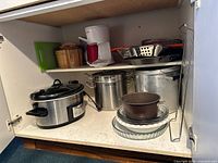 Wide view inside cupboard showing all items including Crock-Pot, coffee maker, pots, colanders, glass dishes, bamboo container, and cutting board