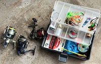 Overview of small plastic tackle box open with organized compartments holding colorful fishing lures, fishing line, and other small fishing accessories, alongside three spinning fishing reels on concrete floor.