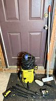 Full view of the Karcher pressure washer with power cord coiled on top, black and yellow spray gun, spray wands, and long brush visible.