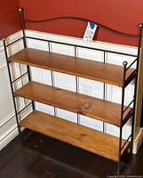 Full view of the bookshelf showing three maple wood shelves and the wrought iron frame with brass finials, placed against red and white wall.