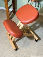 Ergonomic kneeling chair showing orange cushioned seat and knee rest on natural wood frame with caster wheels.