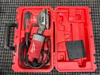 Milwaukee 1/4-sheet palm sander inside its red carrying case, showing the power cord coiled next to the tool, and a black accessory piece or battery holder inside the case.