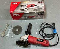 Full lot overview: SKIL angle grinder with box, two wrenches, auxiliary handle, washer, and power cord arranged on a gray floor.