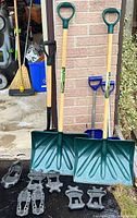 Two snow shovels standing upright near a brick wall, showing wooden handles with green plastic D-shaped grips and wide green plastic scoops, with several black rubber shoe grips laid on the ground in front.
