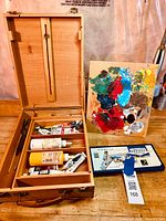 Open wooden artist paint box showing multiple paint tubes and bottles in shelving compartments alongside paint palette and boxed watercolor set.