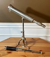Full view of the vintage metal telescope mounted on an adjustable tripod stand with the captains spy glass laid horizontally on the wooden surface.
