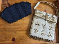 Photo showing both purses on wooden surface: cream beaded fringe purse and black textured clutch