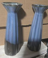 Two blue glazed bud vases shown from the front and side angles against a cardboard background.