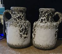 Two textured ceramic jugs with mottled black and white glaze and handles shown from front on a wooden surface.