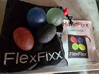 Set of four textured oval hand exercisers in red, blue, green, and black, shown on top of a black FlexFixx branded bag, alongside the HandFixx user guide.