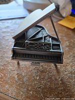 Front angled view of pewter grand piano figurine with open lid, showing keyboard, music stand, and piano legs.
