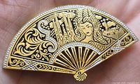 Close-up image of the fan-shaped brooch showing intricate gold and black enamel design with a female figure and floral patterns.