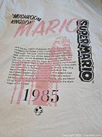 Front view of white Uniqlo T-shirt showing 1985 Super Mario themed pink and black print including text 'Mushroom Kingdom', 'Super Mario', and story text.