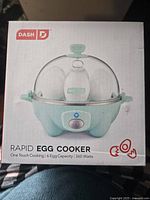 Front of the box showing Dash Rapid Egg Cooker with blue base, clear dome lid, one-touch control button, 6 egg capacity and 360 watts power.