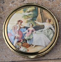 Top view of vintage compact mirror showing painted pastoral family picnic scene on lid.