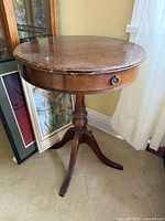 Full view of the round American Empire style pedestal side table, showing wood finish and drawer with brass handle.