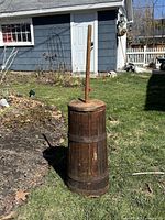 Full view of tall wooden butter churn standing on grass outside near garden and blue house