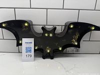 Black metal bat-shaped marquee light with glowing yellow LED lights embedded in wings and body shown from the front on a white tile background.