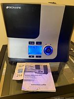 Photo showing the Bionaire BUL9100 humidifier with digital display illuminated and included instruction leaflet on a black glass surface.