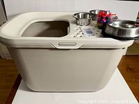 Photo of beige covered cat litter box with top entry hole and three stainless steel bowls plus a cat toy on the lid.