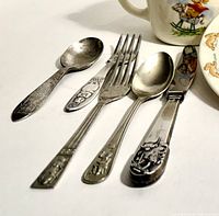 Five children's flatware pieces arranged on a white surface with detailed character motifs and ornate handles.
