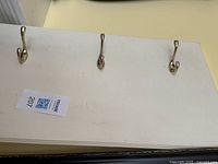 Close-up of three brass hooks mounted on the cream-colored wood panel, showing wear and some surface scratches on the panel surface.