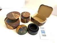 Four assorted handmade jewellery boxes on white background. Two round leather boxes with brown and gold leaf embossed lids, one rectangular leather box open showing plain interior, one round wooden box with painted floral lid.