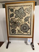 Full front view of the vintage embroidered fireplace screen showing the entire rectangular embroidery panel with stylized floral motifs within a plain wood frame with feet for standing.