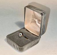 Solitaire-style faux diamond ring displayed inside a grey velvet ring box with cream interior lining.