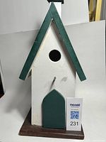 Front view of a decorative folk art birdhouse painted white and dark green with a circular entry hole and door accent on a removable dark brown base.