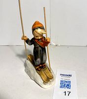 Front view of the Goebel M.J. Hummel little boy skier figurine showing the figure standing on skis with wooden skewers as poles and the orange hat and scarf detail.