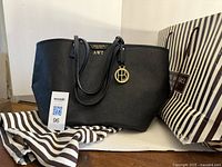 Full view of the black Henri Bendel large tote bag with leather crossgrain texture, gold medallion charm, and handles. Striped bag and shopping bag behind.