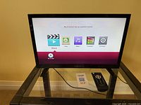 Front view of the Insignia 19-inch LED television powered on, showing its menu with options for movie, photo, music, premium, and settings. A remote control is placed next to the TV on a glass stand.