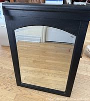 Full view of the large black wooden mirror standing upright on a hardwood floor showing the black decorative crown and beveled mirror glass.