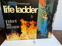 Original box packaging for American LaFrance Life Ladder Instant Fire Escape showing branding and an example use scenario of a woman escaping via the ladder from a second-story window with fire below.