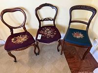 Front view of the three mismatched Victorian wooden parlour chairs with needlepoint seats showing floral embroidery designs in burgundy and dark blue backgrounds.