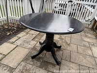 Photo showing round wooden table painted black, fully opened with round top, outdoors on paved surface.