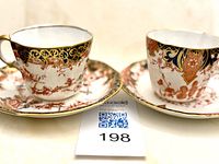 Two Royal Crown Derby teacups with their respective saucers displayed next to each other, showing different yet complementary floral and scroll patterns with gold accents.