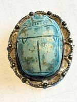 Close-up image of the glass scarab beetle showing detailed carving and patina on a silver or silver-toned mount with rope work and ball embellishments.
