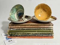 Two mismatched porcelain teacup and saucer sets resting atop a stack of vintage English books with gilt titles visible on spines.