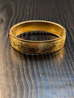 Gold-toned floral engraved bracelet lying on a wooden surface showing detailed pattern and polished inner surface.