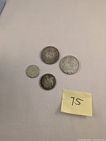 Overhead photo showing all four coins including 1967 Bob Cat Quarter, two Silver Dollars, and 50 cent coin with a note '75'.