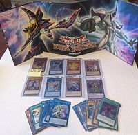 Photo showing the front of the double sided deluxe Duel Power game board with artwork of Dark Magician and a dragon, foregrounded by 17 Yu-Gi-Oh! cards arranged in rows.